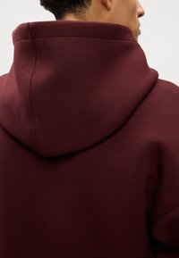 Burgundská mikina s veľkou, jemnou kapucňou, hladkým povrchom a viditeľnými švami na krku na zadnej strane.