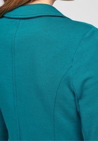 Vêtement bleu sarcelle avec une texture lisse, doté d'un col classique et de coutures subtiles le long du dos, conçu pour une silhouette ajustée.