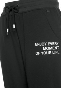 Schwarze Jogginghose aus weichem Baumwollmaterial, mit einem Kordelzugbund und weißem gesticktem Text am linken Bein. Leicht tapered geschnitten.