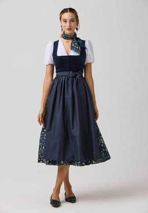 Frau trägt ein marineblaues Midikleid mit weißen Puffärmeln, einem passenden Schal und schwarzen Ballerinas und steht vor einem schlichten Hintergrund.