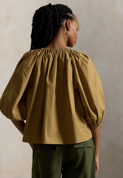 Μουσταρδί βαμβακερό μπλουζάκι με μανίκια-μπαλόνι και ελαστικό ντεκολτέ. Συνδυασμένο με ελαιχρωματικά παντελόνια, με χαλαρή, oversized εφαρμογή.