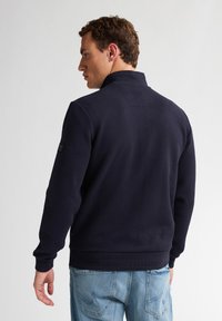 Granatowy sweter z wysokim kołnierzem, teksturowanym diamentowym wzorem, prążkowanymi mankietami i dołem. Nosi go osoba w jasnoniebieskich dżinsach, widziana z tyłu.