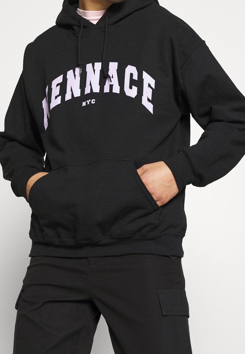 Personne portant un sweat à capuche noir avec le texte « MENNACE NYC » en grandes lettres blanches, les mains dans la poche avant, associé à un pantalon cargo noir.