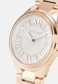 Hodinky Michael Kors s růžovou zlatou kovovou páskou a kulatým bílým číselníkem s římskými číslicemi. Texturované detaily lemují sklo.