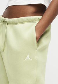 Άτομο με σκουρόχρωμο δέρμα που φοράει ανοιχτό πράσινο παντελόνι φόρμας με λευκό λογότυπο Jumpman, χέρι στη τσέπη και λευκό cropped top.