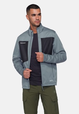 Mann trägt eine grau-schwarze Mammut-Jacke über einem schwarzen Shirt und olivgrünen Cargo-Hosen, richtet den Reißverschluss der Jacke, schaut nach rechts.