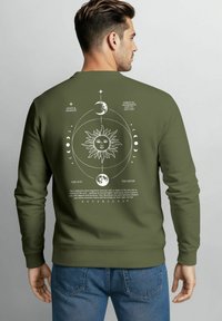 Olive green sweatshirt featuring a circular graphic of the sun and moon, surrounded by text and celestial symbols on the back.