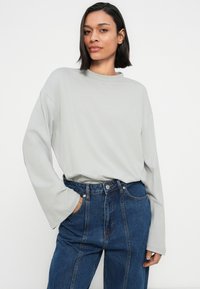 Ανοιχτό γκρι μακρυμάνικο πουκάμισο με ψηλό λαιμό, άνετη εφαρμογή και ραμμένες ώμες. Συνδυάζεται με ψηλόμεσα μπλε τζιν παντελόνια.