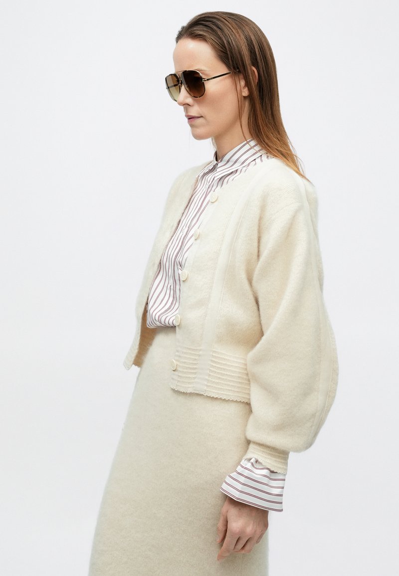 Woman wearing oversized beige cardigan and matching skirt over striped blouse with ruffled cuffs, accessorized with large brown sunglasses.