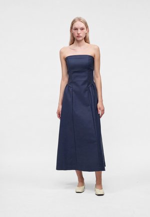 Een vrouw staat en draagt een donkerblauwe strapless midi-jurk en crèmekleurige slip-on schoenen tegen een effen witte achtergrond.
