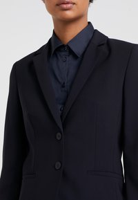 Blazer marine à coupe ajustée, doté d'un revers cranté, de deux boutons noirs et de poches latérales, porté sur une chemise boutonnée assortie.