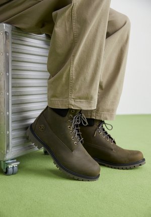 Bottes chelsea en cuir marron avec des bouts ronds, des lacets gris, des œillets renforcés et des semelles en caoutchouc texturé, assorties à un pantalon beige clair.