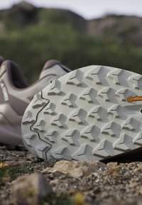 Podeszwa buta trekkingowego z głębokim, wzorzystym bieżnikiem na skalistej ziemi i rozmytym zielonym naturalnym tle.