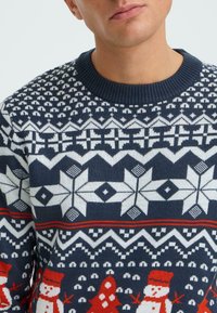 Knit sweater in navy and white featuring snowflake, geometric, and snowman patterns; ribbed crew neck, soft texture, and colorful accents.