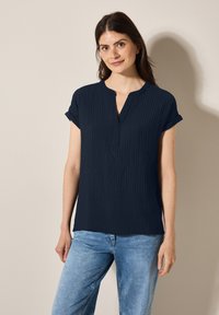 Maglietta blu navy a maniche corte con scollo a V, caratterizzata da un motivo a coste testurizzato. Il materiale sembra leggero e informale. Abbinata a jeans in denim chiaro.
