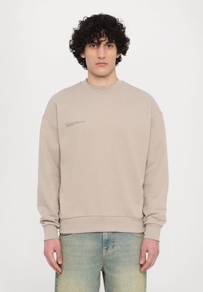 Beige crewneck sweatshirt with long sleeves and ribbed cuffs, featuring minimal text detail on the chest, paired with light blue jeans.