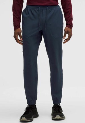 Mann trägt marineblaue Jogginghosen mit elastischen Bündchen, ein bordeauxrotes Langarmshirt und schwarze Sportschuhe, steht vor einem schlichten Hintergrund.