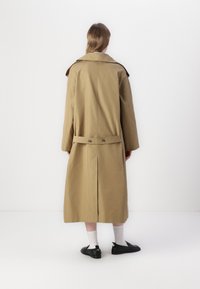 Khaki trenchcoat med bred krage, lange ermer og belte i midjen. Har et bakventil og glatt stoff, akkompagnert av sorte sko.