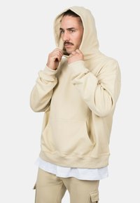Beige hoodie with a kangaroo pocket, ribbed cuffs, and a drawstring hood, worn over a white shirt, featuring a relaxed fit.