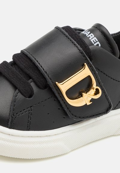 Schwarzer Lederschuh mit Klettverschluss und goldenen Hardware-Details, weißer Gummisohle und strukturierten Stoffschnürsenkeln. Geeignet für Freizeitkleidung.