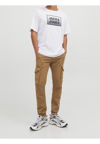 Weißes T-Shirt mit schwarzem "JACK & JONES"-Logo, kombiniert mit beigen Cargo-Hosen mit Seitentaschen. Fußbekleidung: sportliche Sneakers in Schwarz und Weiß.