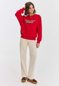 Czerwony sweter z zielonym napisem "mucho amor", luźny krój, ściągacze przy mankietach i u dołu. Zestawiony z beżowymi spodniami i brązowymi butami na sznurowadła.