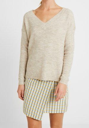 Pull beige côtelé col en V associé à une mini-jupe asymétrique à carreaux jaune, bleu et blanc sur une personne debout.