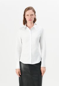 Camicia bianca a maniche lunghe con colletto a punta, chiusura a bottoni, design aderente e texture liscia, abbinata a una gonna in pelle nera.