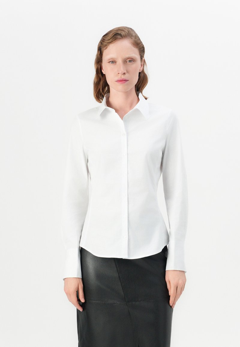Camicia bianca a maniche lunghe con colletto a punta, chiusura a bottoni, design aderente e texture liscia, abbinata a una gonna in pelle nera.