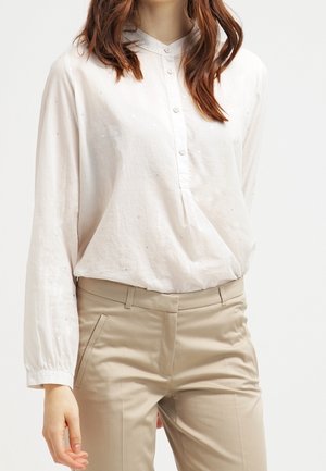 Femme portant une chemise blanche à manches longues boutonnée, rentrée dans un pantalon beige sur mesure, debout avec une main détendue le long du corps.