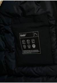 Vnitřní část černé péřové bundy s etiketou obsahující pokyny k údržbě a vlastnosti produktu, včetně ikon pro vodoodpudivost a prodyšnost.