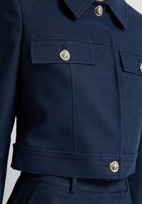 Námořnická modrá zkrácená bunda vyrobená z texturované tkaniny, s dvěma náprsními kapsami, zlatými kovovými akcenty a ostrým designem límce.