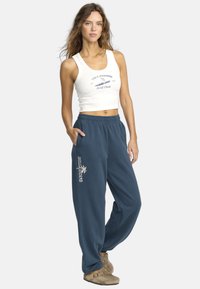 Vit croppad tanktop med blått tryck, kombinerad med lösa blåa joggingbyxor med palmträdets logotyp. Modellen står med en hand i fickan.