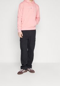 Osoba oblečená v růžové mikině Tommy Jeans, černých cargo kalhotách a hnědých teniskách Adidas s bílými pruhy, stojící na jednobarevném pozadí.