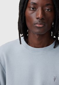 Felpa azzurra con collo tondo, polsini a costine e un discreto logo ricamato sul petto. Texture liscia e design casual.