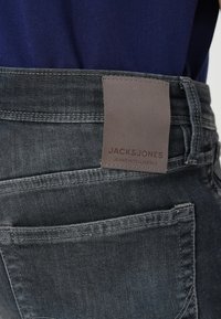 Lähikuva tummanharmaan denimfarkun takataskusta, jossa on ruskea nahkainen merkki, jossa lukee "JACK & JONES JEANS INTELLIGENCE", ja tummansininen paita.