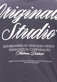 Šedé bavlněné tričko s bílým potiskem. Design zahrnuje "Originální studio" a další branding a informace o založení níže.