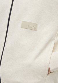 Vêtement crème zippé avec un patch rectangulaire portant l'inscription BOSS sur la poitrine gauche et une main dans la poche latérale.