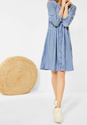 Femme en robe chemise à rayures bleues et blanches, baskets blanches, debout à côté d'un grand tapis rond tissé sur fond blanc.