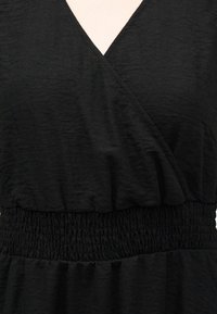 Κοντινό πλάνο μαύρου υφασμάτινου φορέματος με ελαστική μέση με πιέτες και λεπτομέρεια μπούστου τύπου περιτύλιξης σε σχήμα V.