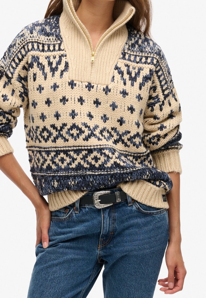 Maglione a maglia con base crema, caratterizzato da motivi geometrici navy, colletto con mezza cerniera e polsini a costine. Indossato sopra jeans a vita alta.