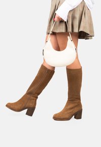 Botas altas de ante marrón con tacón cuadrado y cremallera lateral, combinadas con un bolso de cuero blanco con un diseño curvado y asa corta.