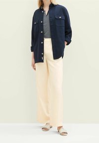 Chemise bleu marine à boutons avec deux poches poitrine, portée sur un top à motifs, associée à un pantalon large crème et des sandales à glissière beiges.