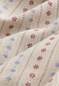 Tissu beige à surface texturée présentant des rangées diagonales de petites fleurs brodées bleu et rouge et des lignes ponctuées assorties.