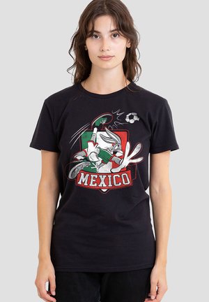 Junge Frau trägt ein schwarzes T-Shirt mit einer Bugs Bunny Grafik, die einen Fußball kickt, und "Mexico" Text vorne vor einem schlichten Hintergrund.