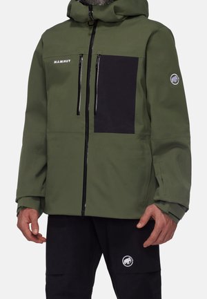 Mies pukeutunut vihreään ja mustaan hupulliseen takkiin, jossa on etuvetoketjut ja Mammutin logot rinnassa ja hihassa, yhdistettynä mustiin housuihin, joissa on Mammutin logo.