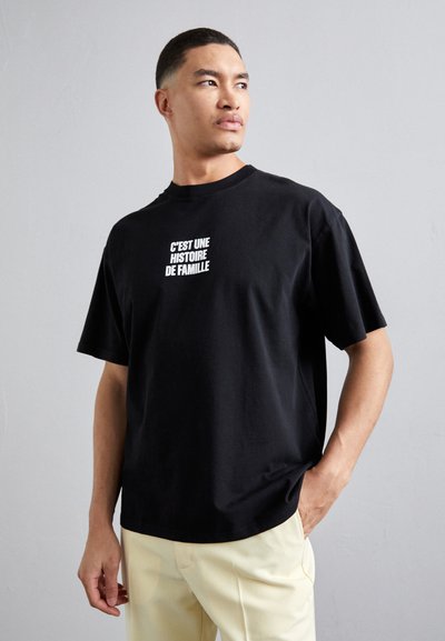 Sort t-shirt i sort bomuld med korte ærmer og rund hals, med hvid tekst der står "C'est une histoire de famille" på forsiden.