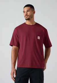 Άνδρας που φοράει μπλούζα μπορντό με κοντά μανίκια και τσέπη στο στήθος, καθώς και μαύρο παντελόνι, στέκεται μπροστά σε απλό ανοιχτό γκρι φόντο.