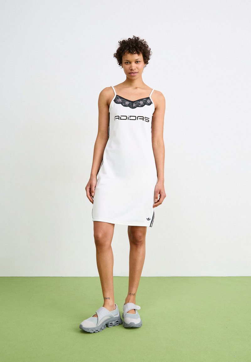 Weißes ärmelloses Kleid mit Spitzenhalsauschnitt und schwarzem "ADIDAS"-Logo. Hat seitliche Streifen und wird mit grauen Sport Schuhen auf einem grünen Boden kombiniert.