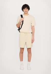T-shirt en coton beige à manches courtes avec une poche poitrine, associé à des shorts beige à cordon de serrage. Baskets blanches et sac à dos noir.